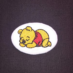 May include: Embroidered patch featuring a sleeping yellow bear wearing a red shirt, set on a white oval. The bear is outlined in black. The patch is likely for clothing or accessories.