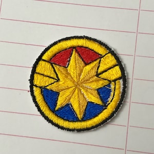 Op de afbeelding: Gestikt patch met het logo van Captain Marvel. Het logo is een gele ster met een rood en blauw centrum, omringd door een gele cirkel met een zwarte omtrek.