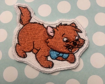 May include: Embroidered patch featuring a cartoon cat in shades of orange and brown, with a blue bow tie. The cat is depicted in a playful pose, outlined with white stitching. Ideal for adding to clothing or accessories.