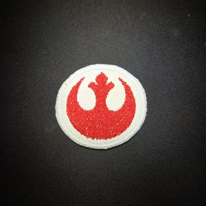May include: A circular embroidered patch featuring a red Rebel Alliance starbird symbol centered on a white background. The design is stitched with a dense thread texture, creating a raised appearance against the fabric surface of the patch.