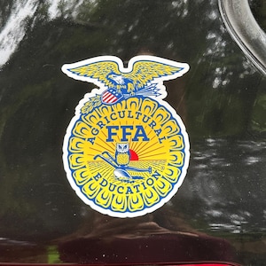 May include: A circular FFA emblem sticker featuring a yellow sun, a blue owl, a plow, and an eagle at the top. The design includes the text Agricultural Education and FFA in blue lettering against a yellow background with a blue border.