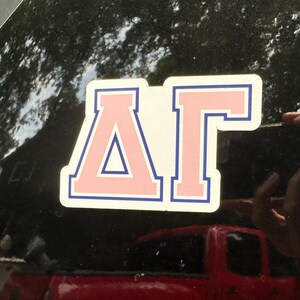 May include: A decal featuring the Greek letters Delta and Gamma in a light pink fill with a dark blue outline. The letters are arranged horizontally and are applied to a smooth, dark, reflective surface, creating a clean and bold visual display.