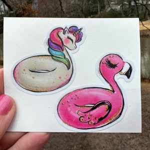 May include: Two stickers featuring whimsical designs. One depicts a unicorn with a rainbow mane and a donut-shaped body. The other is a pink flamingo. Both have gold glitter accents and are on a white background.