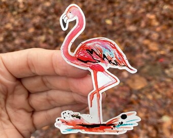 May include: A pink flamingo sticker with watercolor-style paint splatters. The flamingo is standing on one leg with its neck curved in an S-shape. The sticker is on a white background.