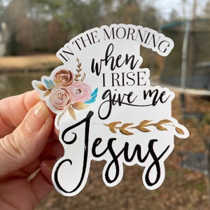 May include: A white sticker with the text "In the morning when I rise give me Jesus" in black script. The sticker has a floral design with gold and pink flowers and gold leaves.