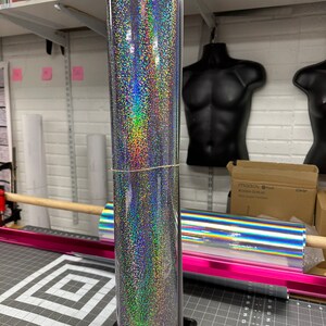 May include: A roll of silver holographic vinyl with a rainbow iridescent finish. The vinyl is rolled up and sitting on a black stand.