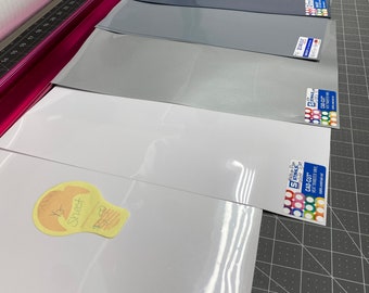 May include: A selection of vinyl sheets in various colors, including white, gray, and black, are arranged on a cutting mat. A roll of clear vinyl is visible in the background. The sheets have product labels and a handwritten note that says "1/2 Sheet".
