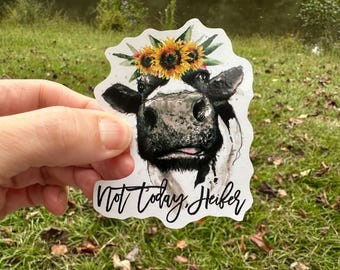 May include: A sticker featuring a watercolor illustration of a cow's face, adorned with a sunflower crown. The cow has a black and white pattern, with the text "Not today, Heifer" in a script font.