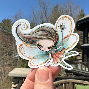 May include: A sticker featuring a whimsical fairy with sparkling gold wings, long brown hair, and a golden wand with a star on top. The fairy is wearing a teal and pink dress with gold glitter accents.