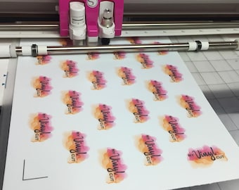 May include: A pink and white vinyl cutting machine precisely cutting a sheet of white paper. The paper is printed with multiple "My Vinyl Cut" logos in black script over watercolor-style orange and pink designs. The machine's blade is in action.
