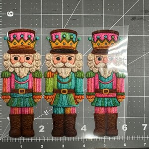 May include: Three colorful nutcracker figures with detailed embroidery. Each has a crown, a white beard, and is dressed in a uniform of pink, green, and brown. The figures are on a gray cutting mat.