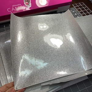 May include: A stack of silver glitter iron-on vinyl sheets. The sheets are on a black cutting mat with white grid lines.