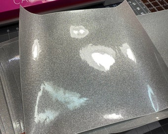 Pode incluir: Uma pilha de folhas de vinil termocolante prateado com glitter. As folhas estão em um tapete de corte preto com linhas de grade brancas.