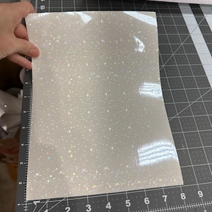 May include: A sheet of white, glittery vinyl with a star pattern. The vinyl is held by a hand and is on a black cutting mat with measurements in inches. A pink paper trimmer is in the background.