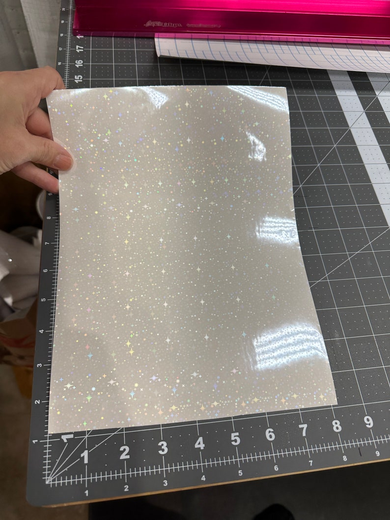 May include: A sheet of white, glittery vinyl with a star pattern. The vinyl is held by a hand and is on a black cutting mat with measurements in inches. A pink paper trimmer is in the background.