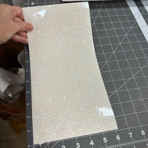 May include: A sheet of white, iridescent vinyl with a sparkling, star-like pattern. The vinyl is held over a black cutting mat with grid lines and measurement markings. A pink cutting machine is visible in the background.