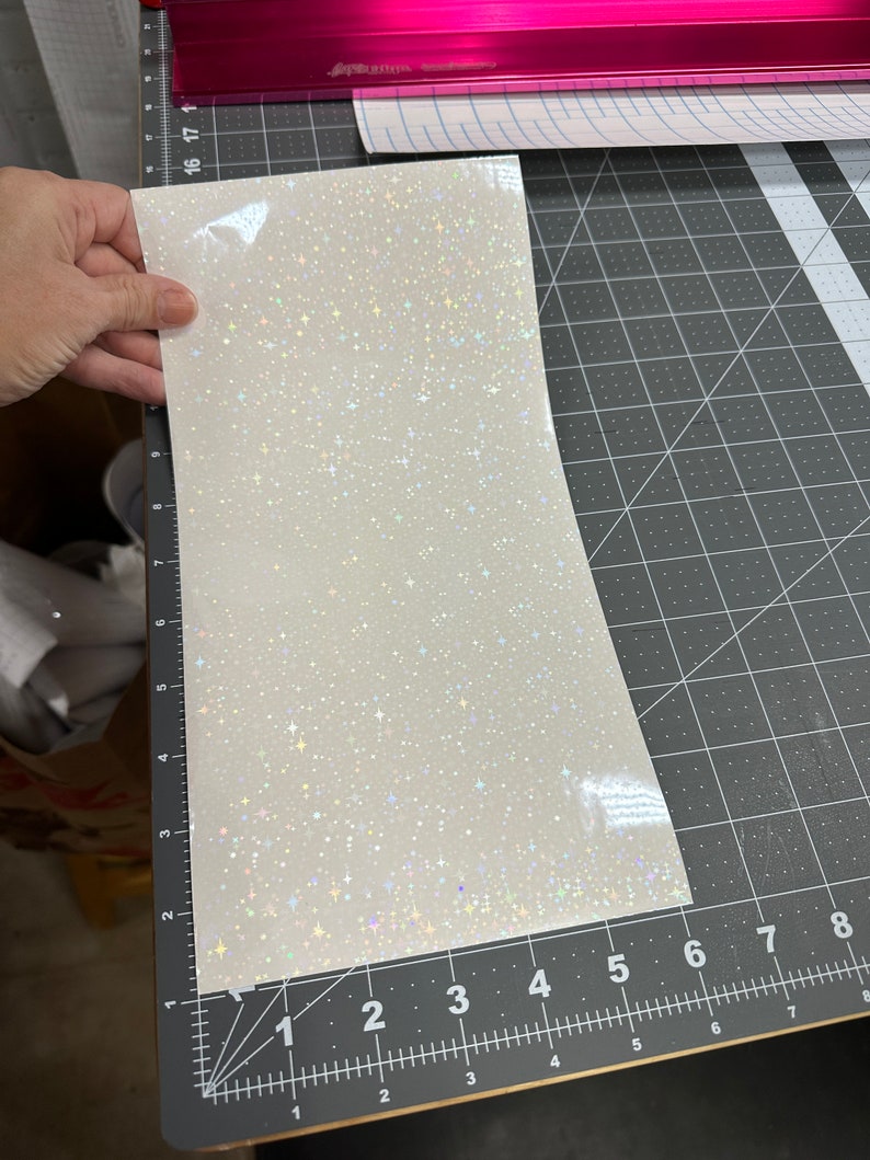 May include: A sheet of white, iridescent vinyl with a sparkling, star-like pattern. The vinyl is held over a black cutting mat with grid lines and measurement markings. A pink cutting machine is visible in the background.