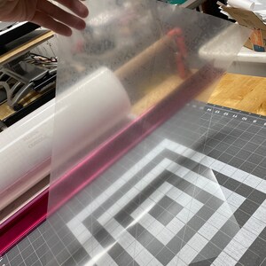 May include: A clear, flexible adhesive transfer sheet with a small rectangular label in the corner featuring the text "CAD-CUT" and "SuperTEK". The sheet is held at an angle over a gray cutting mat with white grid lines and numerical markings.