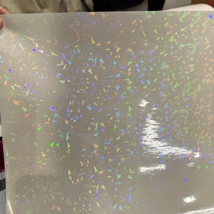 May include: A sheet of iridescent holographic vinyl with a scattered pattern of small, triangular shapes. The vinyl is white with a rainbow sheen.