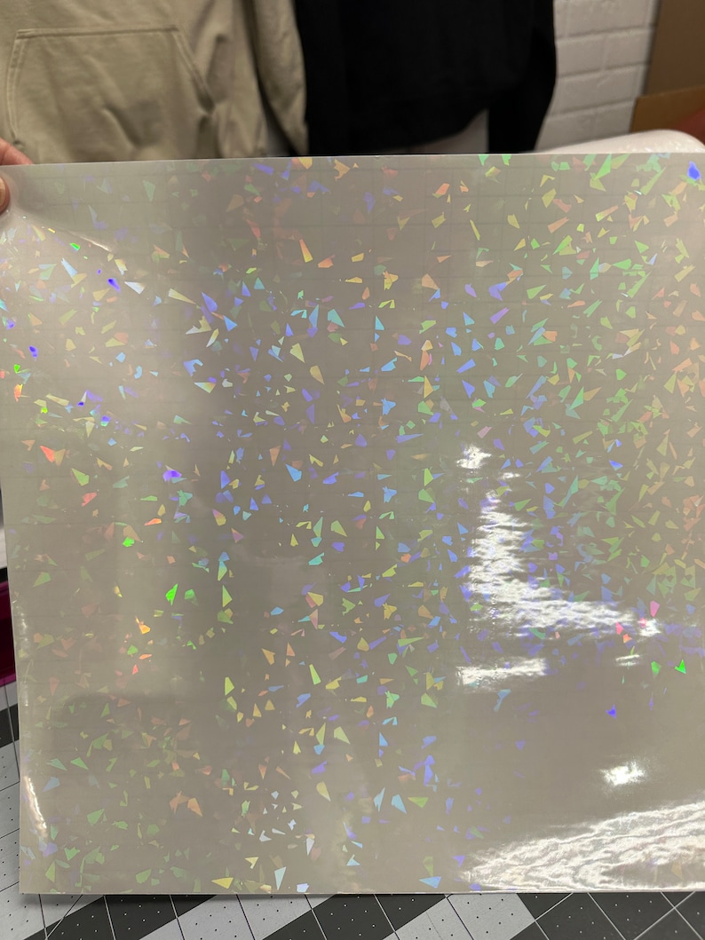 May include: A sheet of iridescent holographic vinyl with a scattered pattern of small, triangular shapes. The vinyl is white with a rainbow sheen.