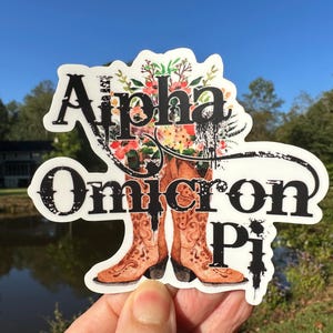 May include: A white sticker with black text that reads "Alpha Omicron Pi" with a floral design and two brown cowboy boots.