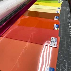 May include: A selection of colorful heat transfer vinyl sheets in shades of coral, orange, yellow, and green. A roll of white vinyl and a pink cutting guide are also visible. The sheets are labeled with product information, including "Fashion Film Coral S4" and "Light Coral 399".