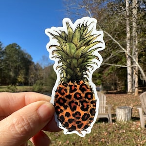 May include: A sticker of a pineapple with a leopard print pattern. The pineapple is drawn in a realistic style with green leaves and a brown body.