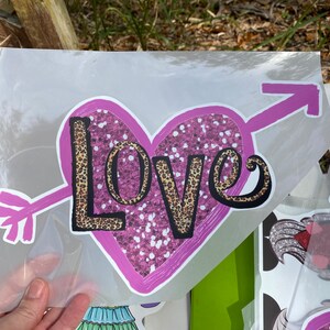Puede incluir: Una pegatina con un coraz&oacute;n rosa atravesado por una flecha rosa. La palabra "LOVE" est&aacute; escrita en el coraz&oacute;n con una fuente de estampado de leopardo con contornos negros. El coraz&oacute;n tiene un interior rosa brillante.
