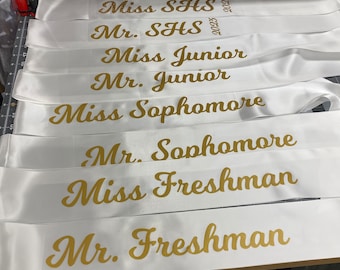 May include: White satin sashes with gold lettering. The sashes read "Miss SHS", "Mr. SHS", "Miss Junior", "Mr. Junior", "Miss Sophomore", "Mr. Sophomore", "Miss Freshman", and "Mr. Freshman".