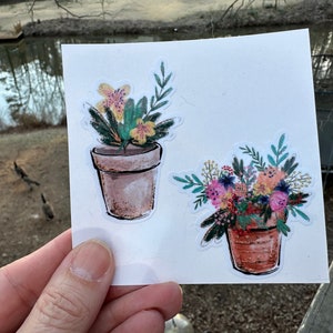 May include: Two watercolor stickers featuring potted flowers. The first sticker shows a yellow flower with green leaves in a brown pot. The second sticker shows a bouquet of pink, orange, and purple flowers in a brown pot.