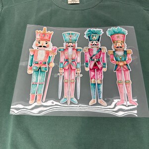 May include: Green crewneck sweatshirt with a heat transfer of four colorful nutcracker figures. The nutcrackers are in pink, teal, and gold, each with a unique hat and holding a sword or staff. The sweatshirt is a solid, muted green.