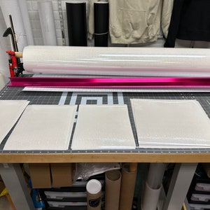 May include: A roll of white glitter vinyl sits above a cutting mat with several cut pieces of the same material. A pink metal guide and cutting tool are also visible. The scene suggests a crafting or DIY project.