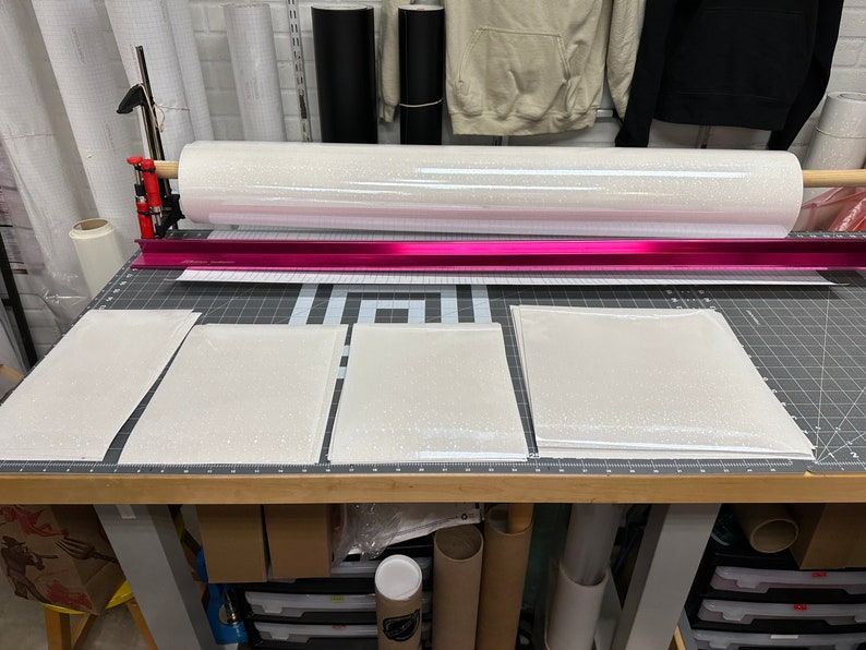 May include: A roll of white glitter vinyl sits above a cutting mat with several cut pieces of the same material. A pink metal guide and cutting tool are also visible. The scene suggests a crafting or DIY project.