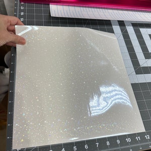 May include: A sheet of white, glittery vinyl with a sparkling, star-like pattern. The vinyl is held up against a ruler marked in inches. A pink cutting guide and other rolls of vinyl are in the background.