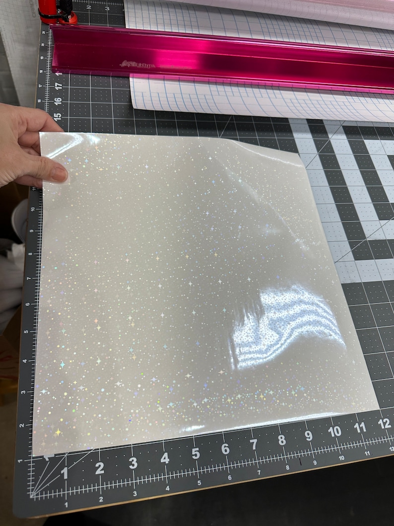 May include: A sheet of white, glittery vinyl with a sparkling, star-like pattern. The vinyl is held up against a ruler marked in inches. A pink cutting guide and other rolls of vinyl are in the background.