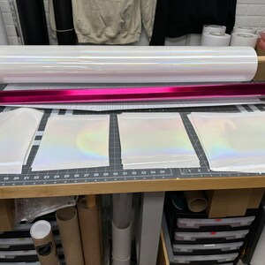 May include: A roll of iridescent white vinyl with a pink core, and three sheets of the same vinyl laid out on a wooden table with a grid pattern.