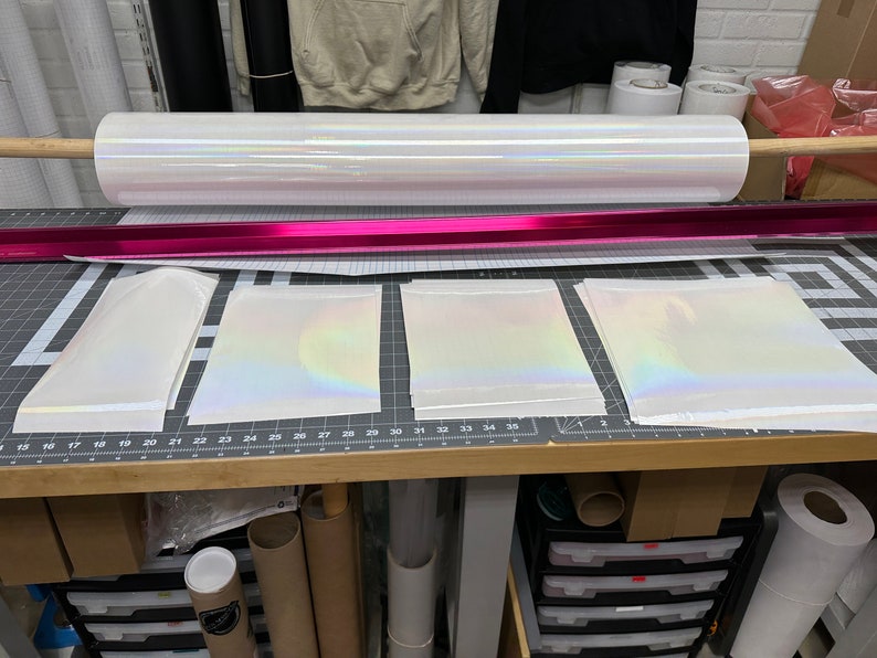 May include: A roll of iridescent white vinyl with a pink core, and three sheets of the same vinyl laid out on a wooden table with a grid pattern.