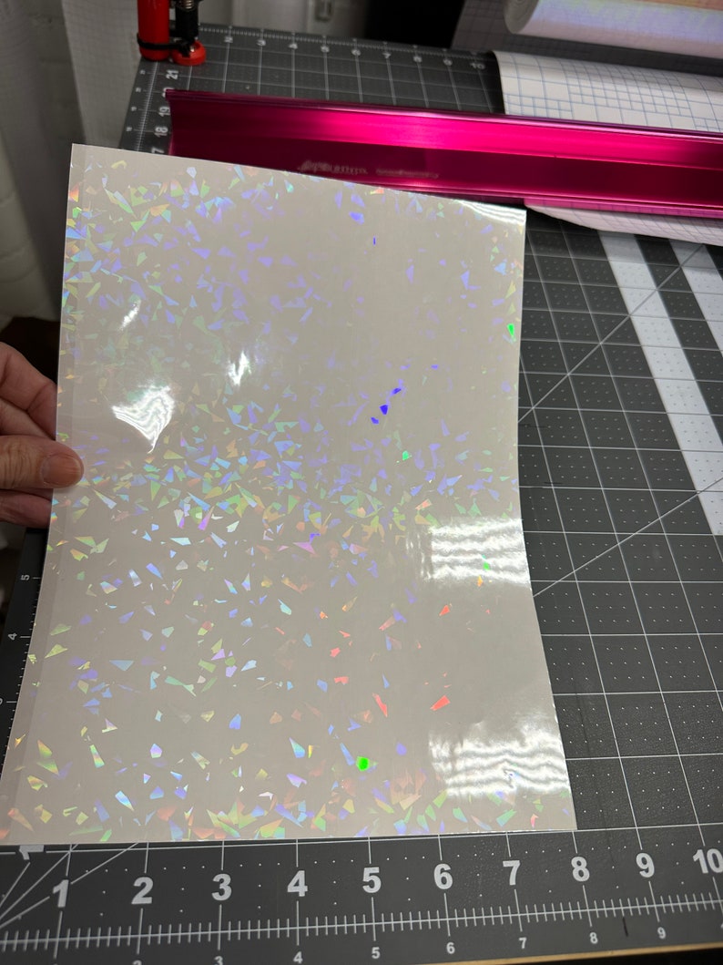 May include: A sheet of iridescent white vinyl with a holographic finish. The vinyl has a scattered pattern of small, iridescent triangles.