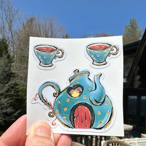 May include: A sticker sheet featuring a whimsical tea set design. The set includes two teacups and a teapot, all in a teal color with gold accents. The teapot has a red door and a yellow glow inside, suggesting a cozy, fairy-tale theme.
