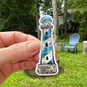 May include: A sticker featuring a lighthouse design. The lighthouse has a blue and white color scheme with gold accents. The top is blue, and the body is white with blue and gold details. The base is a stone-like texture.