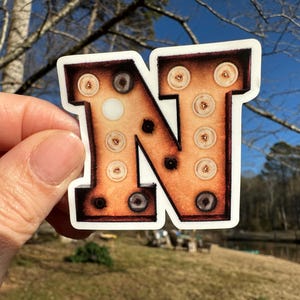 May include: A white sticker with a brown and orange letter N. The letter is made to look like a vintage marquee sign with light bulbs.