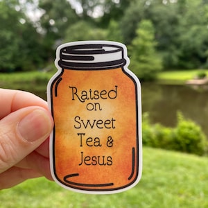 May include: A sticker of a mason jar with a brown and orange background. The jar has the text "Raised on Sweet Tea & Jesus" written on it.