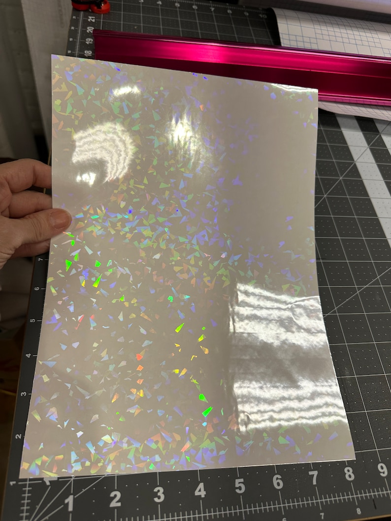 May include: A sheet of iridescent white vinyl with a holographic finish. The vinyl has a scattered pattern of small, iridescent triangles.