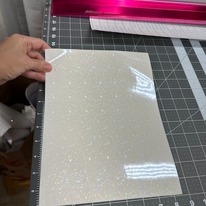 May include: A sheet of white, glittery vinyl with a starry pattern is held over a cutting mat. The vinyl has a shiny, iridescent finish. A pink metal ruler and rolls of vinyl are in the background.