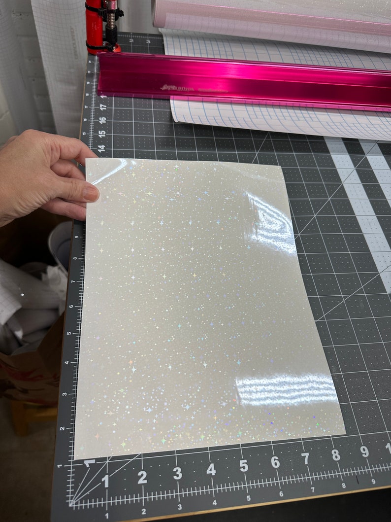 May include: A sheet of white, glittery vinyl with a starry pattern is held over a cutting mat. The vinyl has a shiny, iridescent finish. A pink metal ruler and rolls of vinyl are in the background.