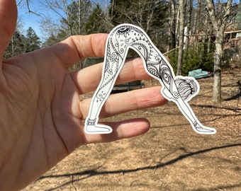 May include: A white sticker featuring a black line drawing of a person in a downward-facing dog yoga pose. The design includes intricate patterns and details on the body, with a focus on the legs and back. The sticker is held in a hand.