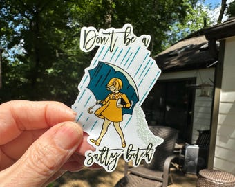 May include: A sticker featuring a cartoon illustration of a person in a yellow dress holding a teal umbrella in the rain. The text reads "Don't be a salty bitch."