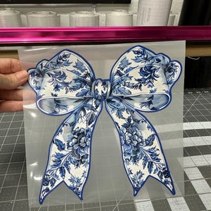 May include: A blue and white floral patterned bow with a white background. The bow is on a clear sheet of transfer paper.