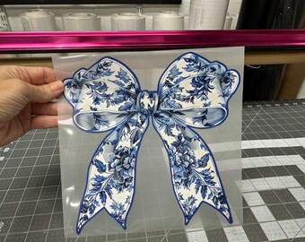 May include: A blue and white floral patterned bow with a white background. The bow is on a clear sheet of transfer paper.