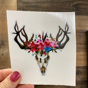 May include: A white sticker with a floral skull design. The skull is surrounded by pink, red, and blue flowers and feathers.